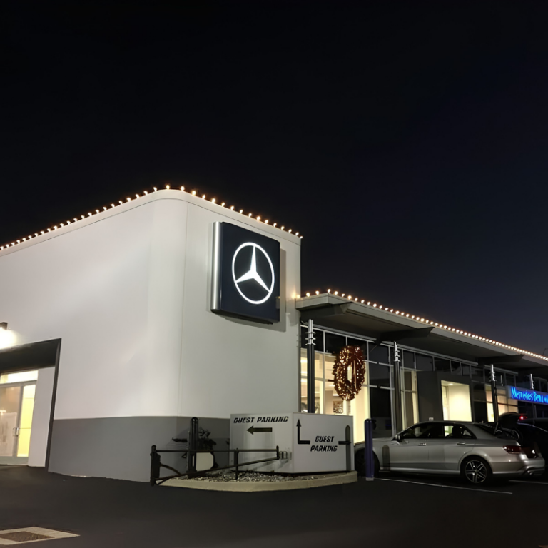 Warm white Christmas lights installed along the rooflines of a car dealership at night, with a lit wreath over the showroom entrance
